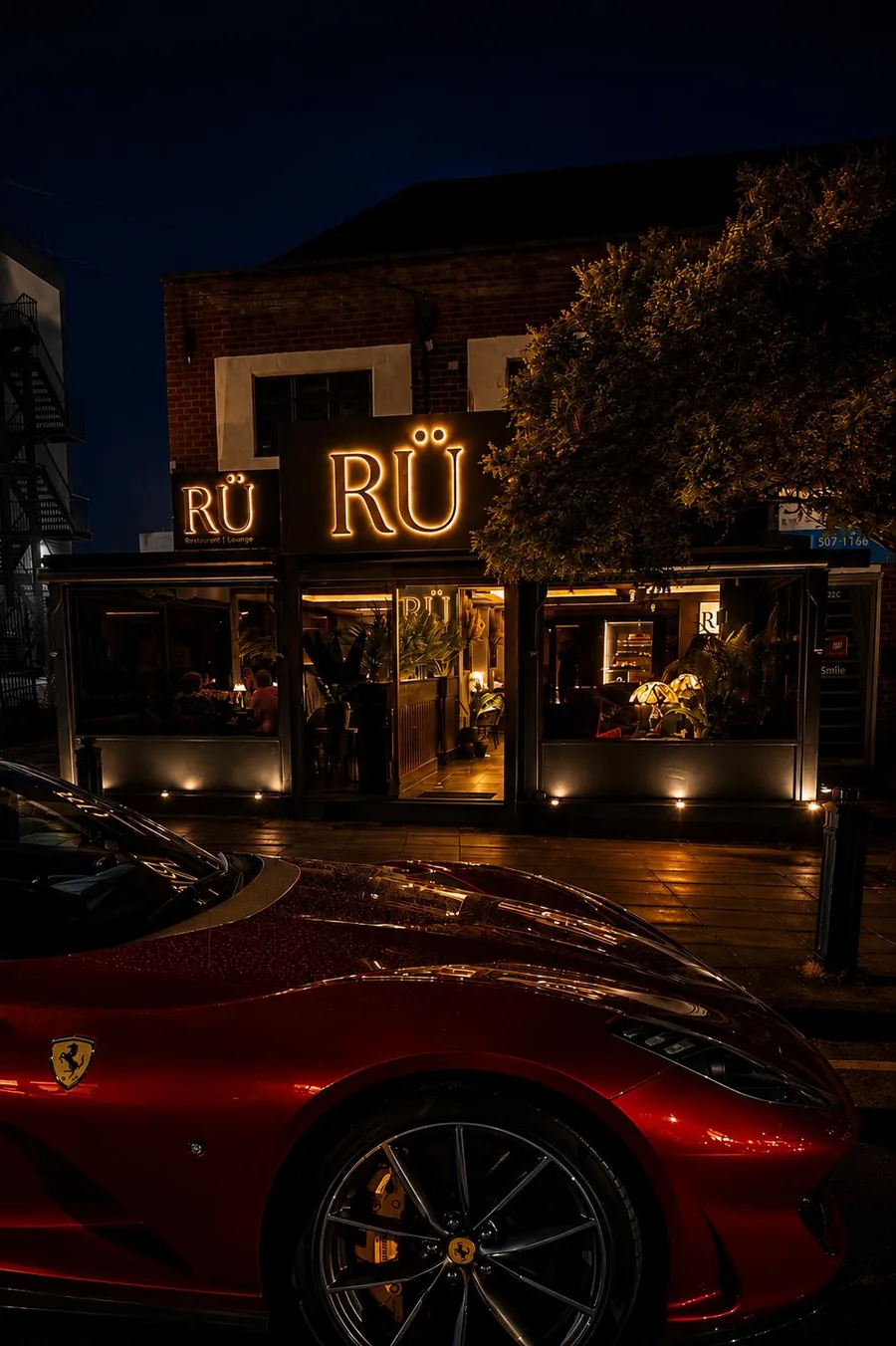 RÜ Restaurant & Lounge lit up at night on Station Road, Cheadle Hulme, with a red Ferrari parked outside