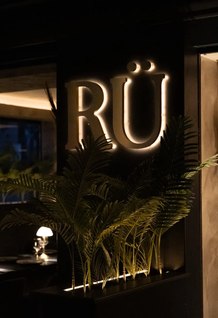 Back-lit RÜ sign framed by palms inside the restaurant